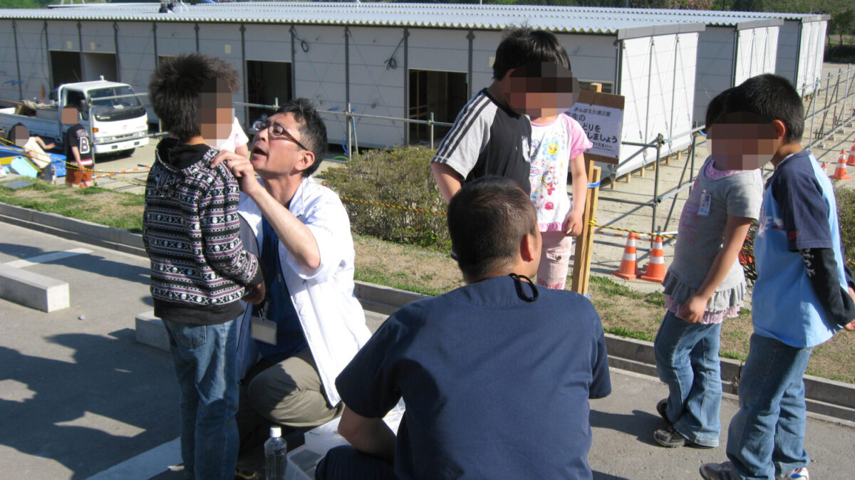 災害時に、屋外で子供たちの歯科検診をする歯科医(白衣の人)(背中が写っている紺色の服の人は太田秀人さん)